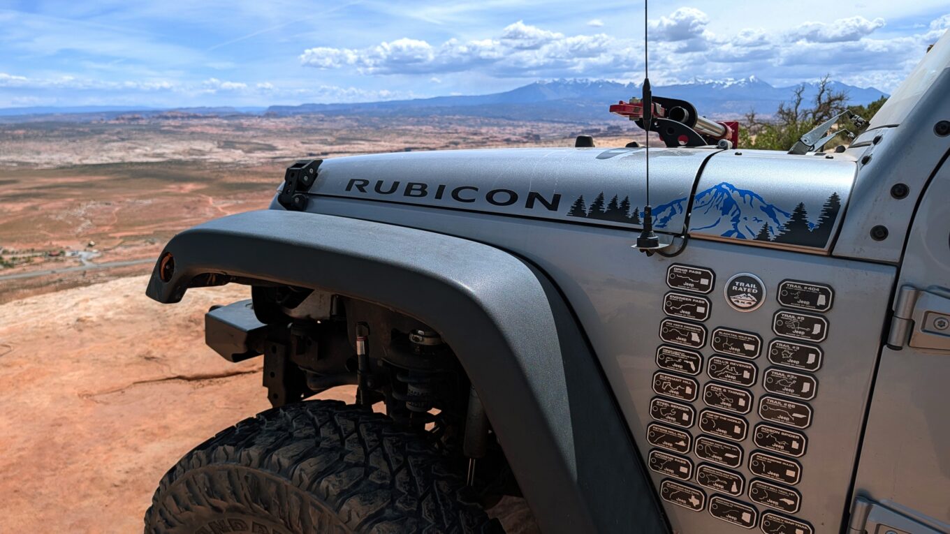 Photo of a silver Rubicon JK Jeep at the edge of the cliff over Moab with the La Sale mountains in the distance. Jeep Badges of Honor visible on the side of the Jeep.